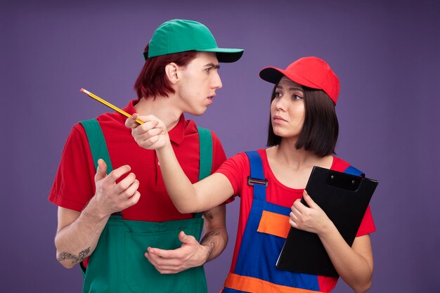Frowning young couple in construction worker uniform and cap girl holding pencil and clipboard looking and pointing at side with pencil guy keeping hand in air looking at girl isolated