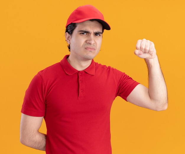 Free photo frowning young delivery man in blue uniform and cap keeping hand behind back looking at front doing knocking gesture isolated on orange wall