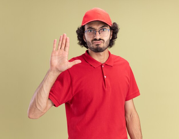 赤い制服を着た眉をひそめている若い配達人とオリーブグリーンの壁に分離された停止ジェスチャーをしている正面を見て眼鏡をかけている帽子