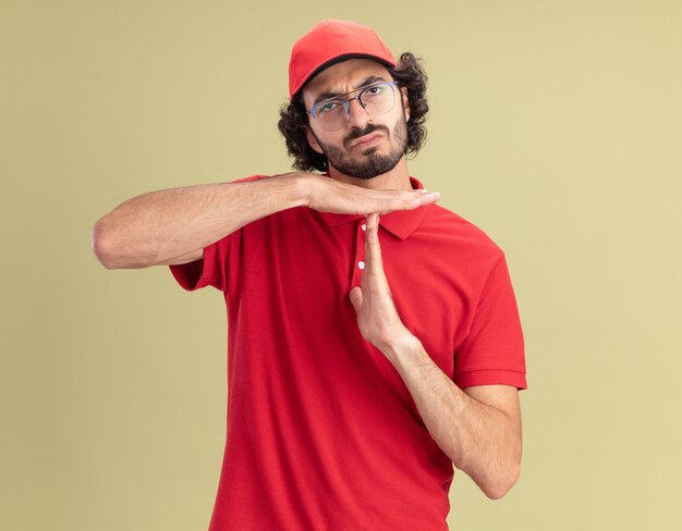 Frowning young delivery man in red uniform and cap wearing glasses looking at front doing timeout gesture isolated on olive green wall