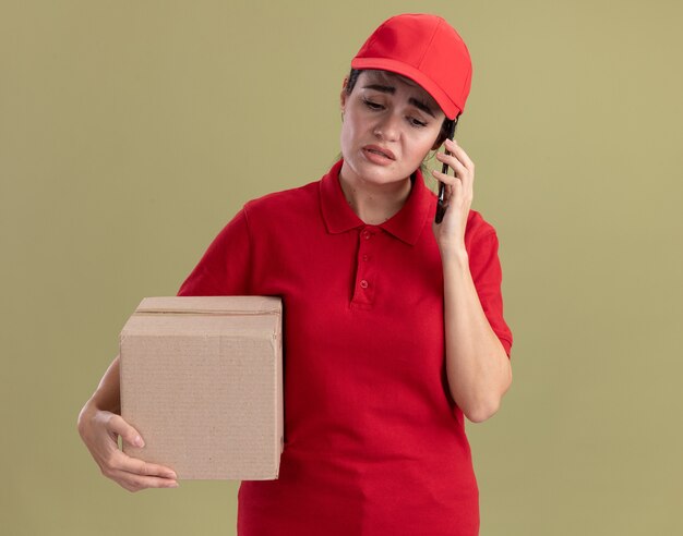 Frowning young delivery woman in uniform and cap holding cardbox talking on phone looking down isolated on olive green wall with copy space