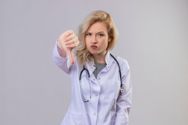 Frowning young doctor wearing stethoscope in medical gown her thumb down on white wall