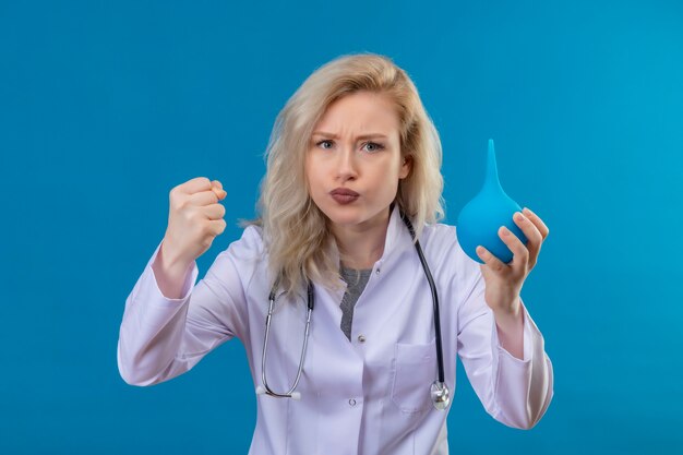Frowning young doctor wearing stethoscope in medical gown holding enema on blue wall