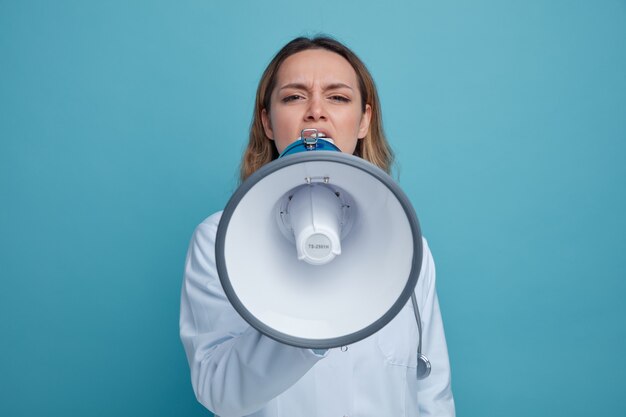 Frowning young female doctor wearing medical robe and stethoscope around neck talking by speaker 