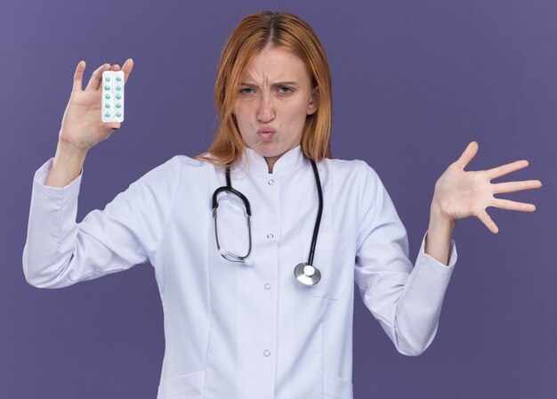 의료 가운과 청진기를 입은 젊은 여성 생강 의사가 보라색 벽에 격리된 입술을 오므린 채 빈 손을 보여주는 전면에 의료 약 팩을 보여주고 있습니다.