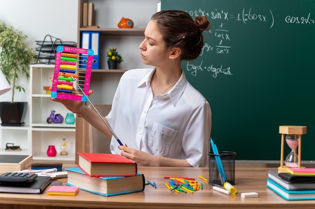 frowning young female math teacher sitting at desk with school supplies holding and looking at abacus pointing at it with pointer stick in classroom