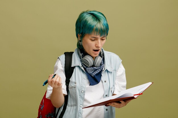 Frowning young female student wearing headphones and bandana on neck and backpack holding pen and open note book looking at note book isolated on olive green background