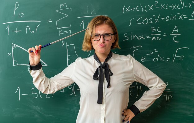 frowning young female teacher standing in front blackboard holding pointer stick on head in classroom