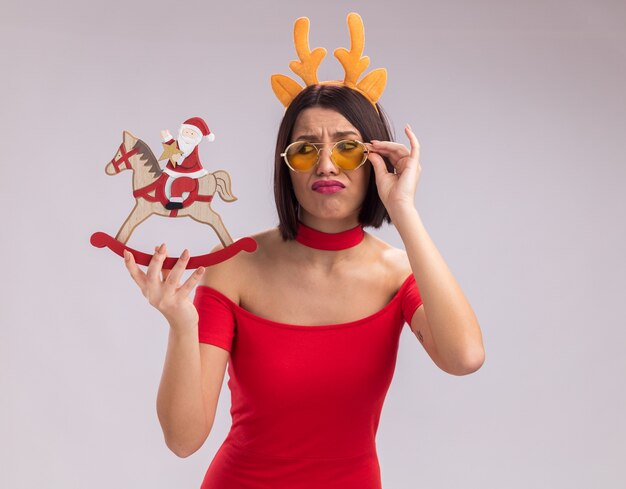 Frowning young girl wearing reindeer antlers headband and glasses holding and looking at santa on rocking horse figurine grabbing glasses isolated on white background