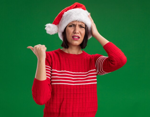 Frowning young girl wearing santa hat  keeping hand on head pointing at side isolated on green wall