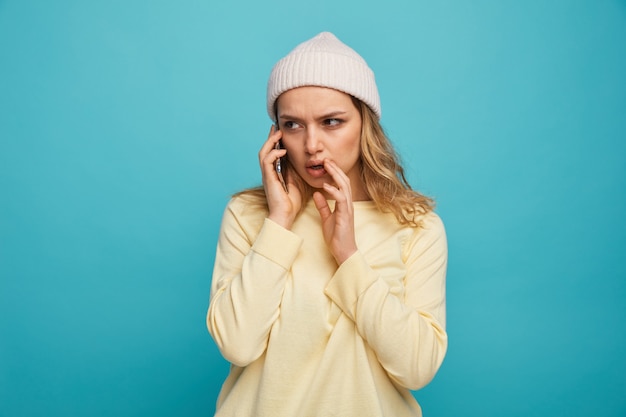 Frowning young girl wearing winter hat talking on phone keeping hand near mouth whispering looking at side 