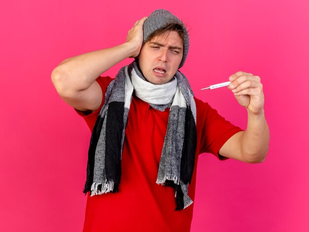 Frowning young handsome blonde ill man wearing winter hat and scarf holding and looking at thermometer touching head isolated on pink wall