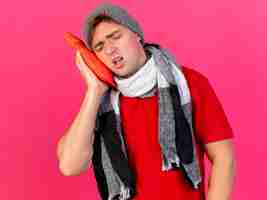 Foto gratuita accigliato giovane uomo malato bello biondo che indossa cappello invernale e sciarpa toccando il viso con una bottiglia di acqua calda con gli occhi chiusi isolato sulla parete rosa