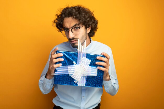 Frowning young handsome caucasian man wearing glasses holding gift pack  isolated on orange wall with copy space