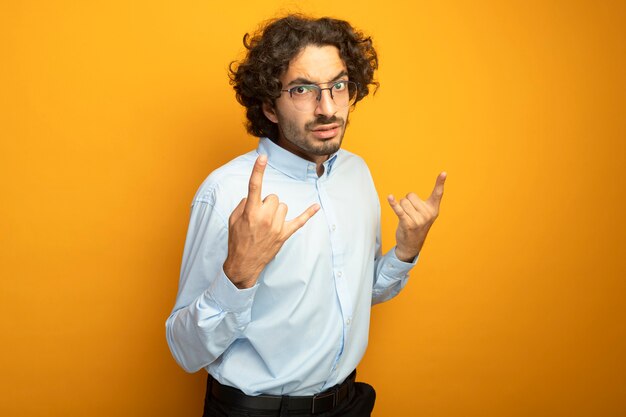 Frowning young handsome caucasian man wearing glasses looking at camera doing rock signs isolated on orange background with copy space