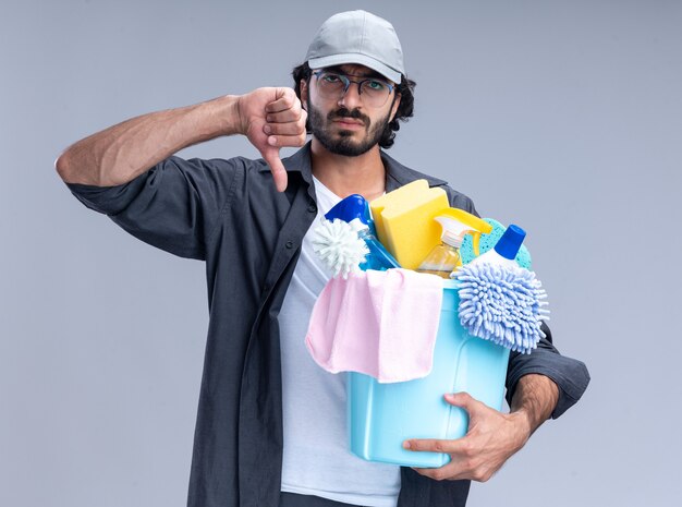 Frowning young handsome cleaning guy wearing t-shirt and cap holding bucket of cleaning tools showing thumb down isolated on white wall