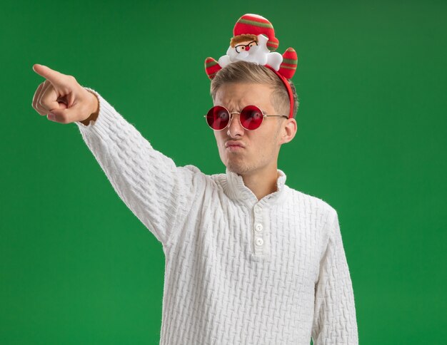 Frowning young handsome guy wearing santa claus headband with glasses looking and pointing at side isolated on green wall