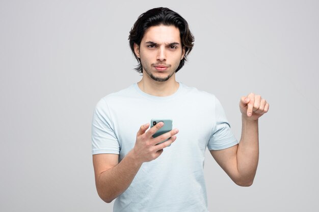 frowning young handsome man holding mobile phone looking at camera pointing down isolated on white background