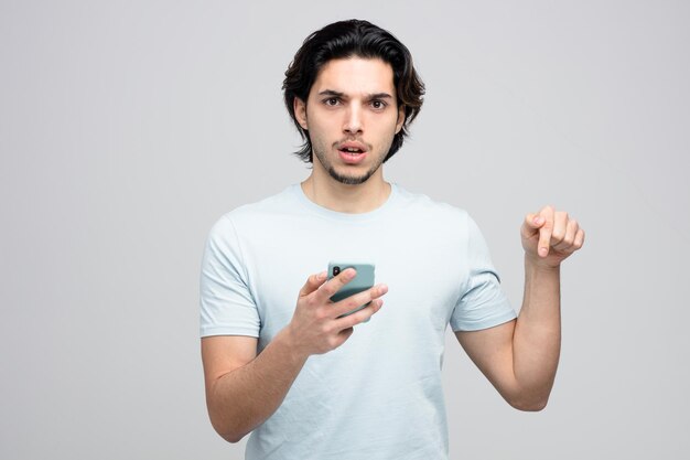 frowning young handsome man holding mobile phone looking at camera pointing down isolated on white background