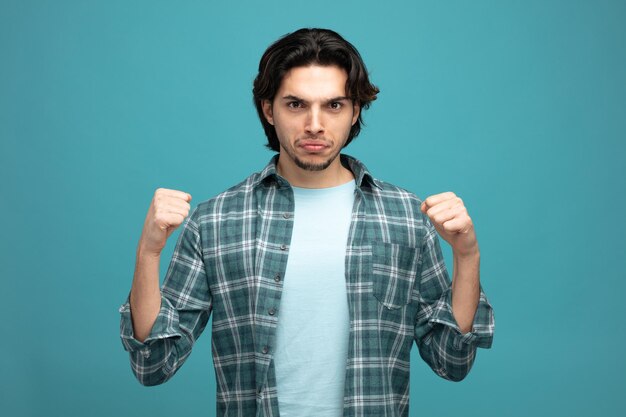 frowning young handsome man looking at camera keeping fists in air isolated on blue background