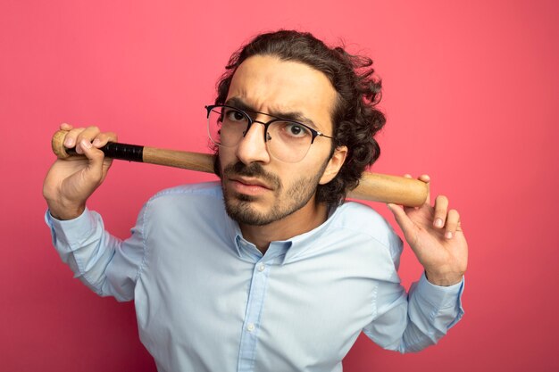 Frowning young handsome man wearing glasses holding baseball bat behind neck looking at front isolated on pink wall