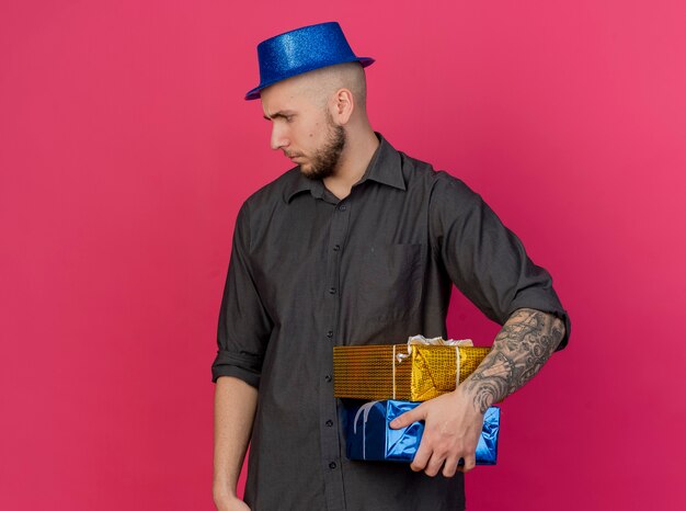 Frowning young handsome party guy wearing party hat holding gift packs looking at side isolated on pink wall