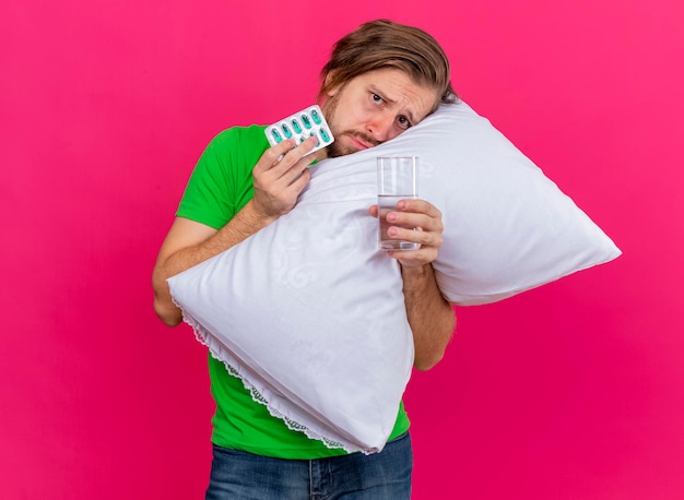 Frowning young handsome slavic ill man hugging pillow looking at front holding pack of capsules and glass of water isolated on pink wall with copy space