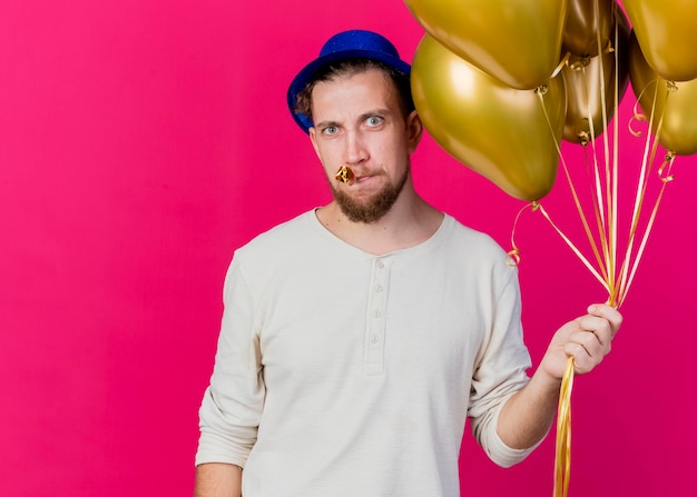 Frowning young handsome slavic party guy wearing party hat holding balloons and party blower in mouth looking at front isolated on pink wall with copy space