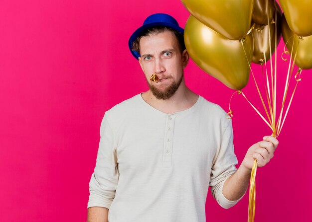 Frowning young handsome slavic party guy wearing party hat holding balloons and party blower in mouth looking at front isolated on pink wall with copy space