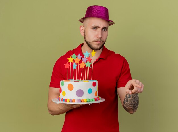 Frowning young handsome slavic party guy wearing party hat holding birthday cake looking and pointing at side isolated on olive green background with copy space