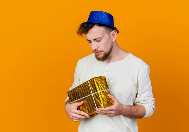 Frowning young handsome slavic party guy wearing party hat holding gift box with closed eyes isolated on orange background with copy space
