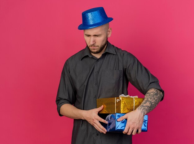 Хмурый молодой красивый славянский тусовщик в партийной шляпе держит подарочные пакеты с закрытыми глазами, изолированными на малиновом фоне с копией пространства