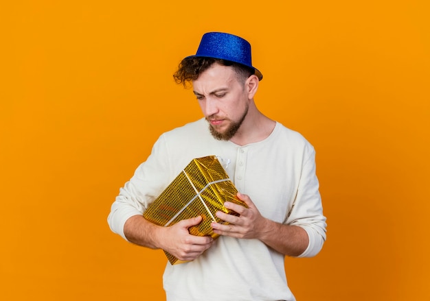 Frowning young handsome slavic party guy wearing party hat looking down holding gift box isolated on orange background with copy space