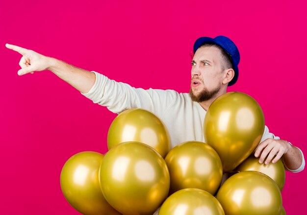 Frowning young handsome slavic party guy wearing party hat standing behind balloons looking and pointing at side isolated on crimson background