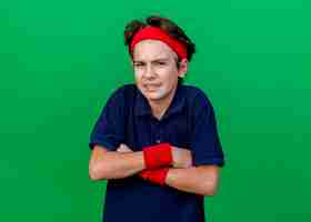 Бесплатное фото Хмурый молодой красивый спортивный мальчик с головной повязкой и браслетами с брекетами, стоя в закрытой позе, глядя вперед, изолированный на зеленой стене с копией пространства