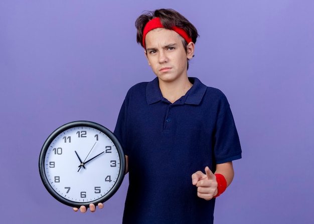 젊은 잘 생긴 스포티 한 소년을 입고 시계를 가리키는 치과 교정기와 팔찌를 착용하고 복사 공간이 보라색 벽에 고립