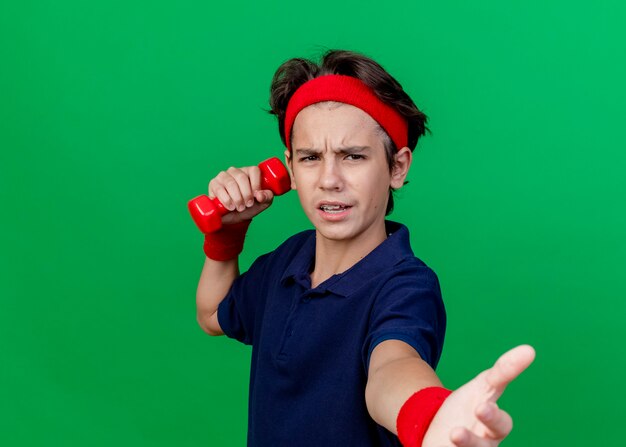 Frowning young handsome sporty boy wearing headband and wristbands with dental braces holding dumbbell  stretching out hand towards isolated on green wall with copy space