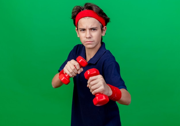 Frowning young handsome sporty boy wearing headband and wristbands with dental braces holding dumbbells doing boxing gesture isolated on green wall with copy space