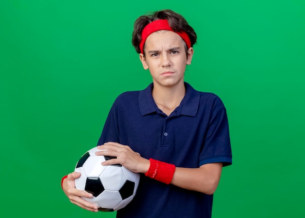 Frowning young handsome sporty boy wearing headband and wristbands with dental braces holding soccer ball looking at front isolated on green wall with copy space