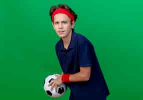 Free photo frowning young handsome sporty boy wearing headband and wristbands with dental braces holding soccer ball looking at front isolated on green wall with copy space