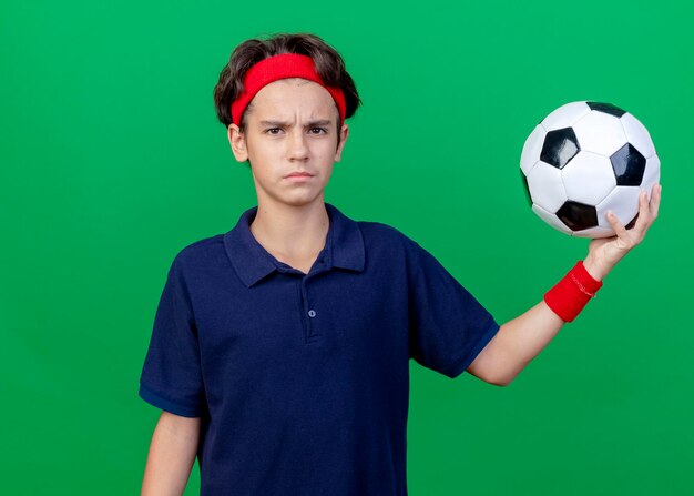 Frowning young handsome sporty boy wearing headband and wristbands with dental braces holding soccer ball looking at front isolated on green wall