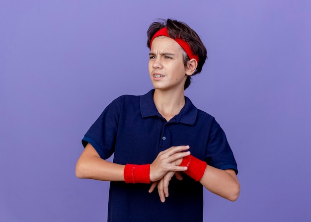 Frowning young handsome sporty boy wearing headband and wristbands with dental braces looking at side putting hand on another one exercising isolated on purple wall with copy space