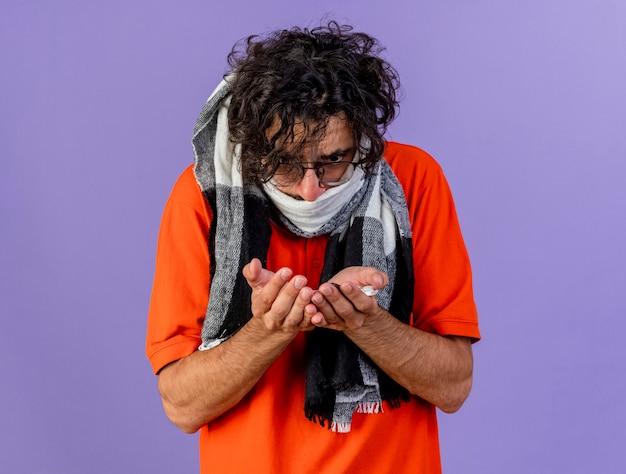 Frowning young ill man wearing glasses and scarf holding and looking at medical pills isolated on purple wall