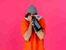 Foto gratuita accigliato giovane uomo malato con gli occhiali inverno cappello e sciarpa che copre la bocca con sciarpa guardando davanti isolato sul muro rosa