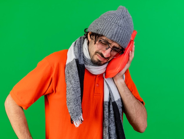 Foto gratuita accigliato giovane uomo malato con gli occhiali inverno cappello e sciarpa che tiene la borsa dell'acqua calda toccando il viso con gli occhi chiusi isolati sulla parete verde