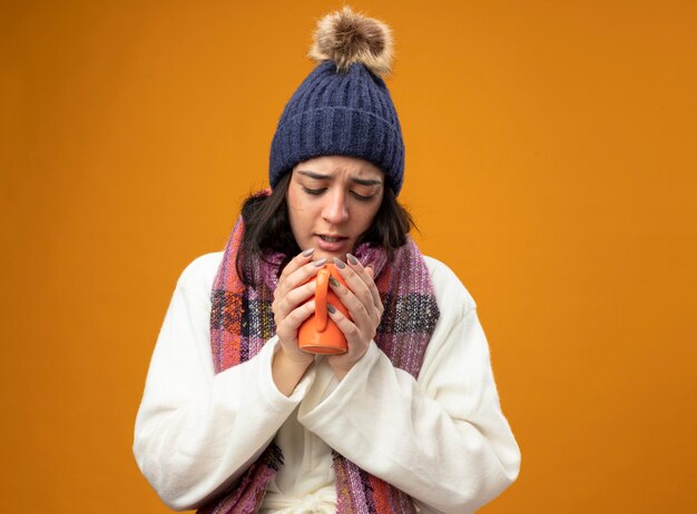 オレンジ色の壁に隔離されたお茶のカップを保持し、見ているローブの冬の帽子とスカーフを身に着けている眉をひそめている若い病気の女性