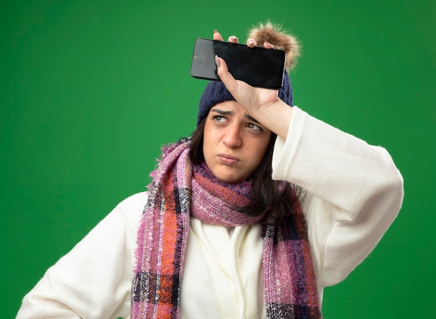 Foto gratuita accigliata giovane donna malata che indossa abito invernale cappello e sciarpa tenendo il telefono cellulare toccando la fronte guardando il lato isolato sulla parete verde