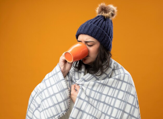 オレンジ色の壁に隔離された目を閉じて格子縞をつかむ格子縞の飲み物のカップに包まれたローブの冬の帽子をかぶった眉をひそめている若い病気の女性
