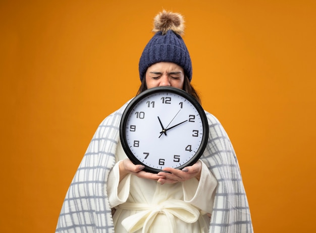 Frowning young ill woman wearing robe winter hat wrapped in plaid holding clock with closed eyes isolated on orange wall