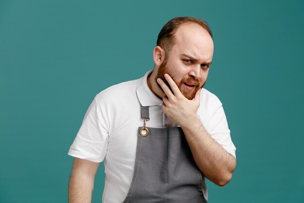 Frowning young male barber wearing white shirt and barber apron looking at camera while keeping hand on chin isolated on blue background